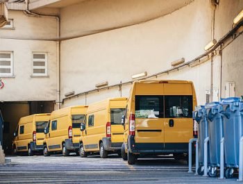 Row of yellow delivery and service van, trucks and cars in front of the entrance of a warehouse distribution logistic plant